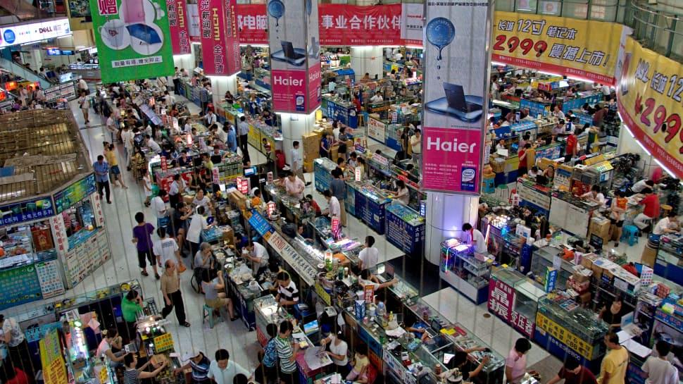 Busy electronics market interior with stalls, shoppers, and Haier ads.