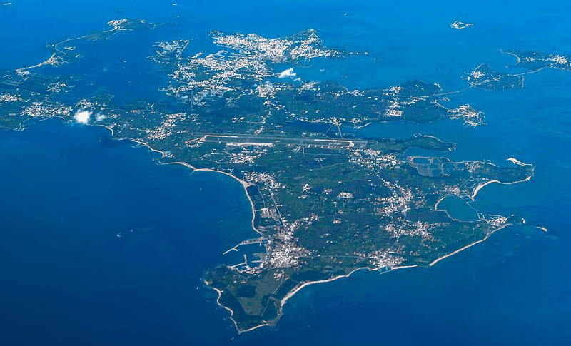 An aerial view of an island surrounded by blue sea water, featuring a winding coastline. The island has multiple towns and residential areas with densely packed buildings, green spaces, and farmland. The center of the island is covered with a significant amount of vegetation, possibly forest or parks. A noticeable airport runway is located at one end of the island, indicating potential air traffic. The overall terrain appears relatively flat with no significant mountains or hills.
