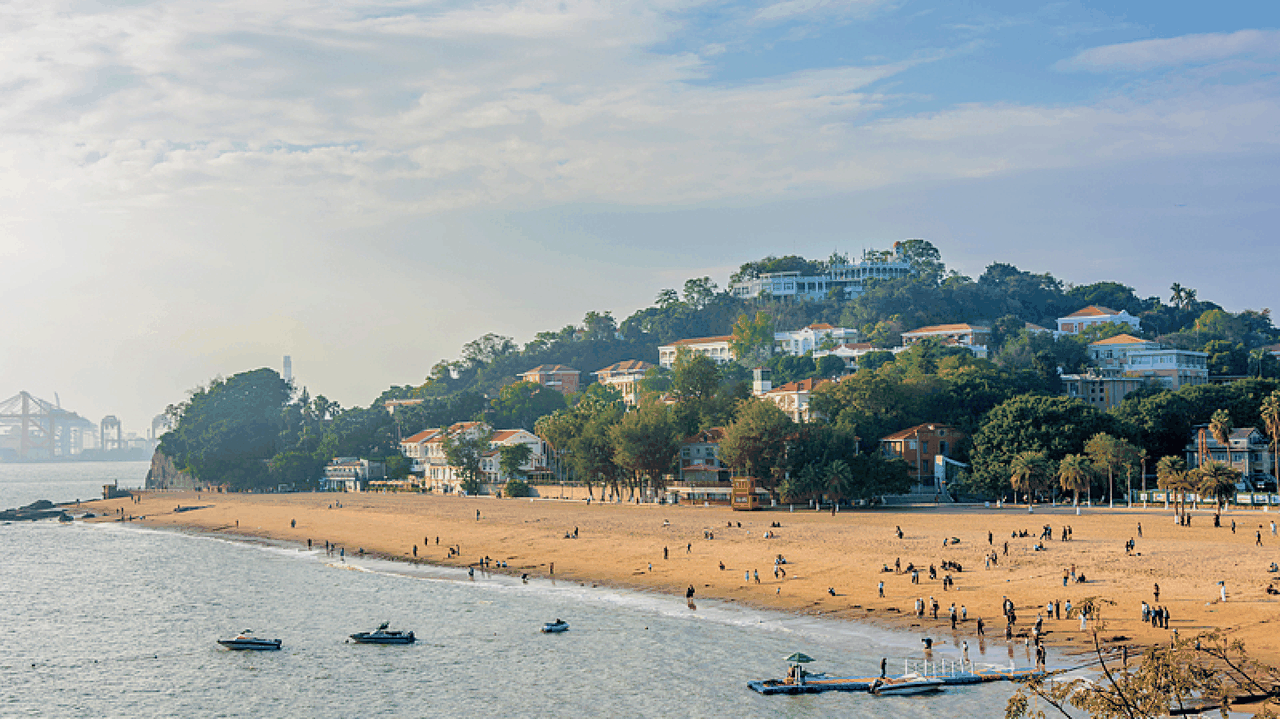 A scenic view of Gulangyu Island's beach, featuring soft golden sand, gentle waves lapping at the shore, and lush greenery in the background.